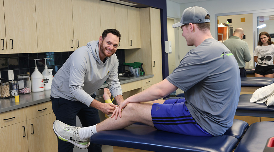 A man in the training facilities smiling at the camera as he works on the leg of a student athlete who's sitting on the exam table