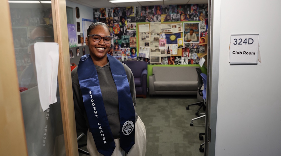 Kayci Resende-Abbott smiles as she stands in the doorway to an on-campus office while wearing a graduation stole that says "student leader"