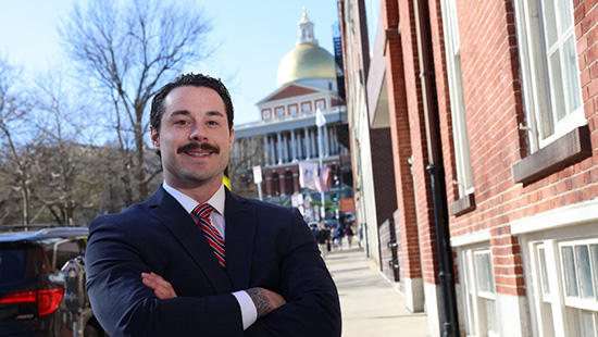 Cody Black stands outside in professional attire, arms crossed, with the Massachusetts State House in the background