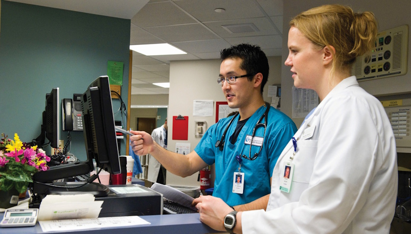 Two clinicians, one in blue scrubs and one wearing a lab coat, stand at a nurses' station in a hospital environment reviewing information on a computer screen