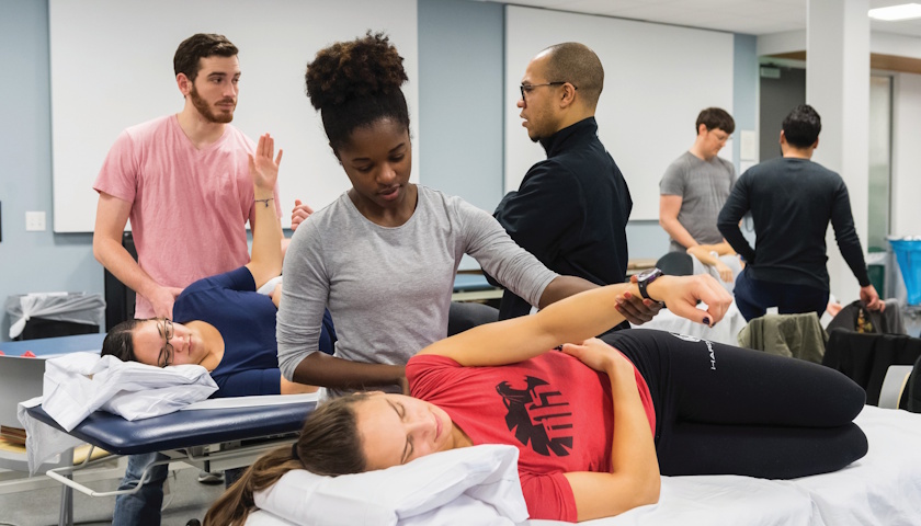 In a physical therapy treatment room, several PT students work with patients. Foreground shows a PT student helping a patient, who is on a table, lift her arm