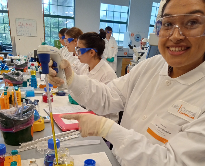 Smiling student at a bench in a lab wearing PPE and a LabCentral lab coat measures liquid in a pipette