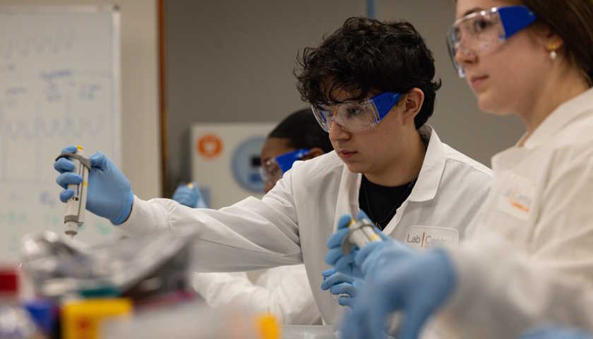 Three students wearing white lab coats and PPE work at a lab bench, while the central figure of the three uses a pipette.