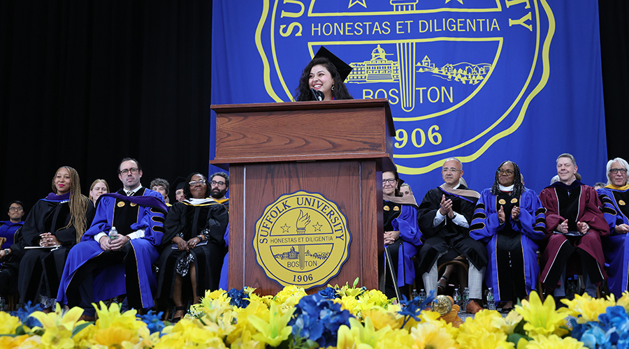 Spriha Paudel smiles at the podium surrounded by Suffolk faculty and administrators on stage at Commencement 2026