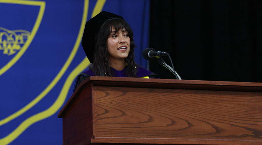 Wearing cap and gown and standing in front of the Suffolk seal, Crystin Mueller addresses fellow graduates at the 2026 Suffolk Law School Commencement