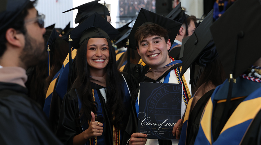 Two Sawyer Business School graduates in caps and gown, smiling and holding up a Commencement program