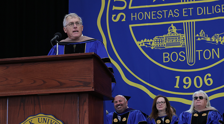 Standing behind a podium with the Suffolk seal behind him, outgoing board chair Robert C. Lamb gives a speech at Commencement