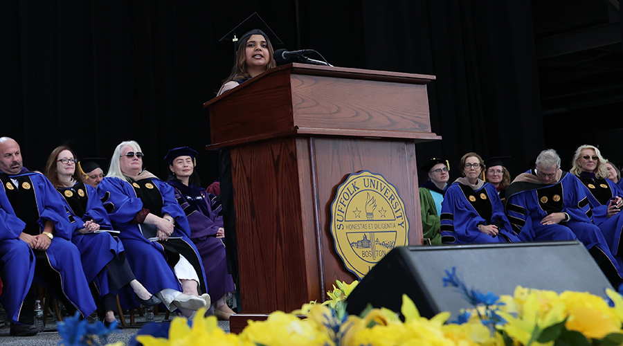 Standing at the podium with faculty behind her and yellow flowers in front of her along the dais, graduate speaker Kavya Sethumadhavan addresses members of the Class of 2026 at the Sawyer Business School Commencement