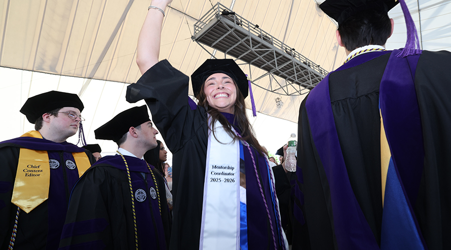 Wearing cap and gown a happy student waves to someone in the crowd ahead of the 2026 Suffolk Law School Commencement