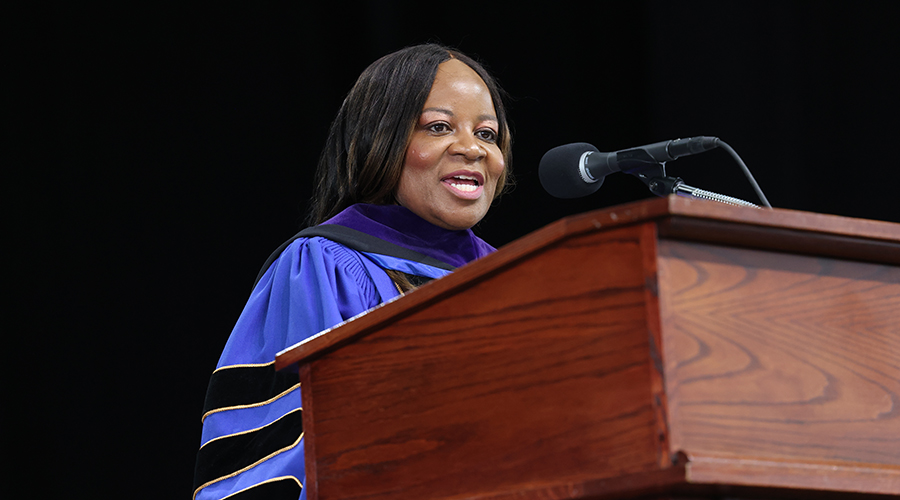 Wearing blue and black regalia, Cheree McAlpine speaks from the podium during the 2026 Suffolk Law School Commencement