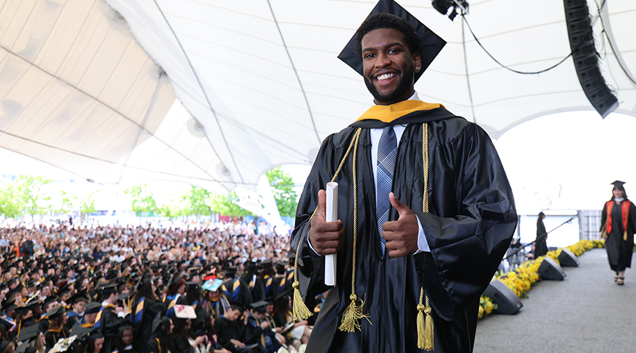 Wearing cap and gown and a colorful stole, a graduate gives a thumbs up with his diploma as he crosses the stage.