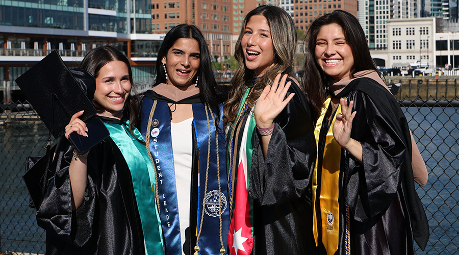Four friends in academic regalia smile and wave in front of the water before the SBS Commencement ceremony begins