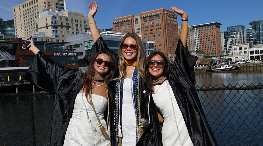 Three friends in academic regalia and sunglasses raise their hands in triumph in front of the water before the SBS Commencement ceremony begins