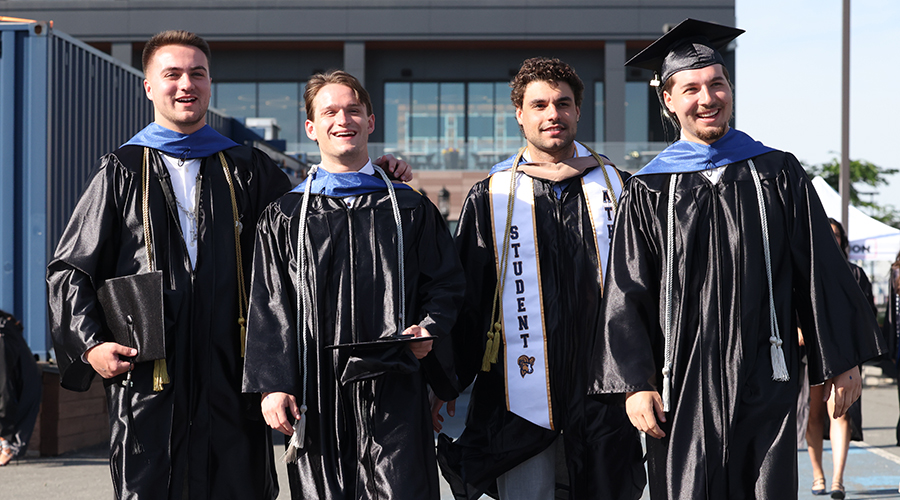 Four friends in academic regalia smile together before the SBS Commencement ceremony begins