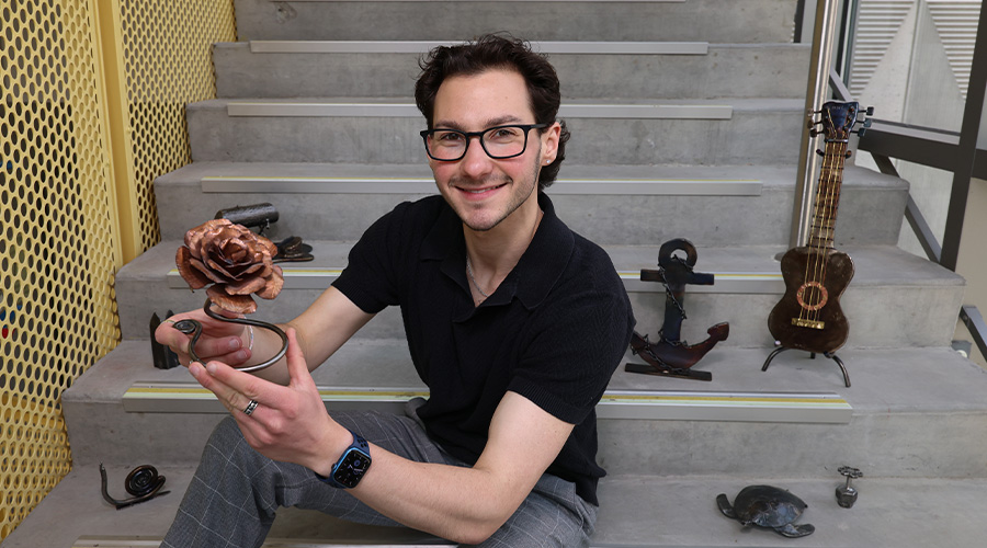 Marcus Frem, Class of 2026, poses on concrete stairs surrounded by his metal sculptures; he's holding his bronze rose scultpure