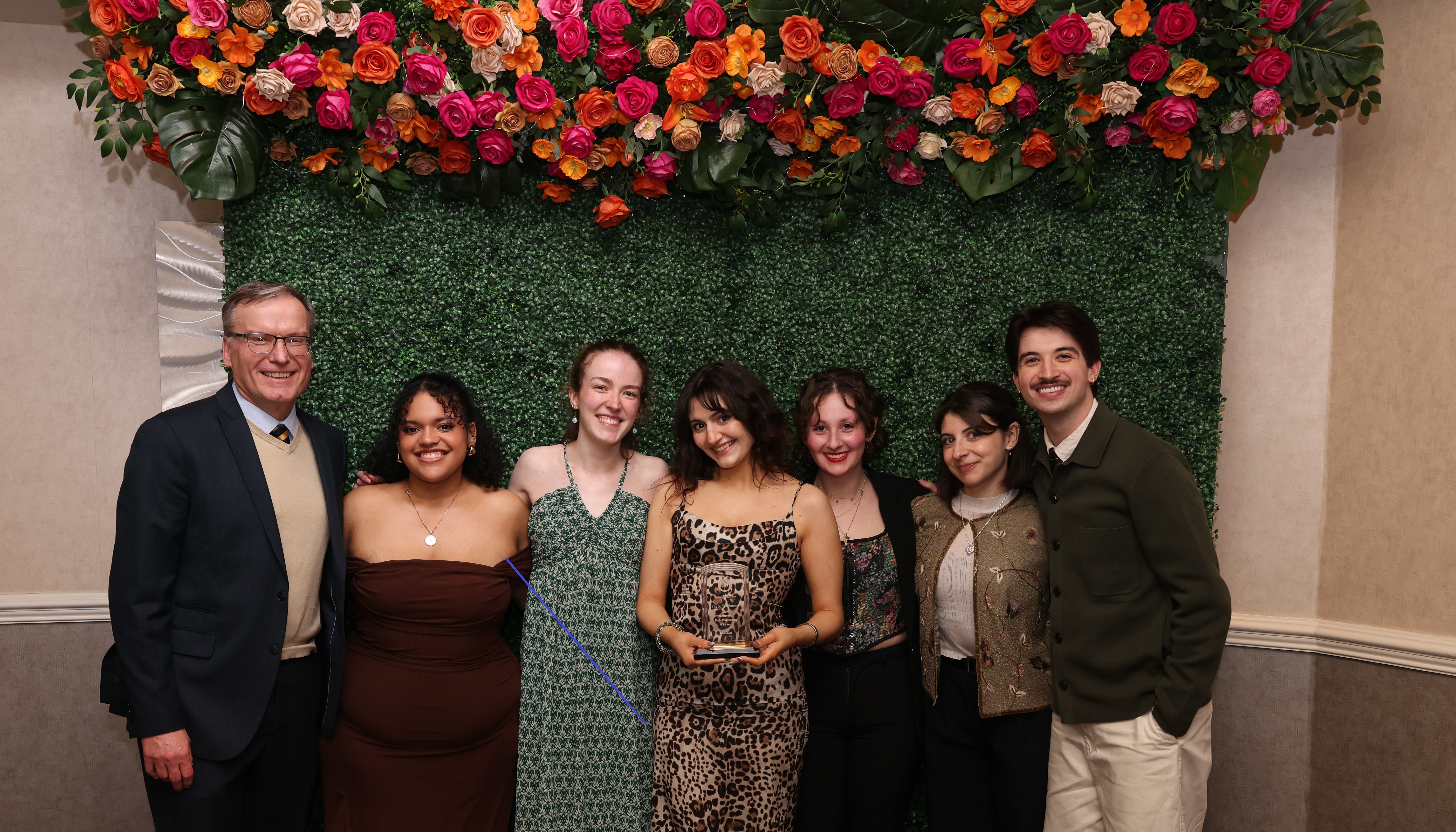 Six Suffolk Journal staffers celebrate their organization's 2026 SGA award alongside advisor Professor Charles St. Amand, posing under a floral canopy