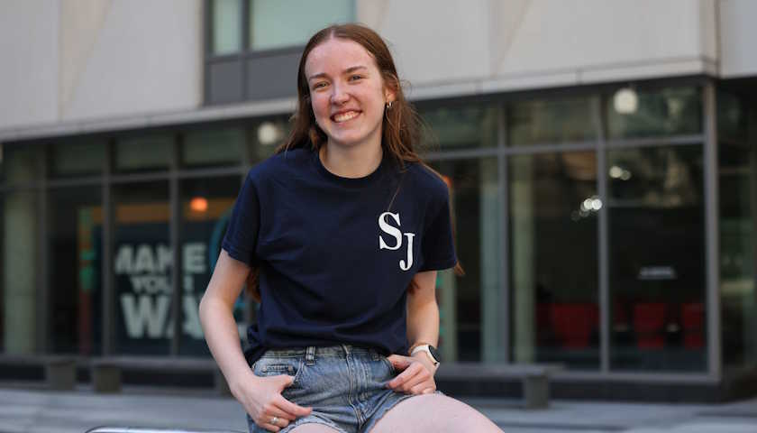 Maren Halpin smiles as she sits outside on the Suffolk campus wearing a Suffolk Journal shirt