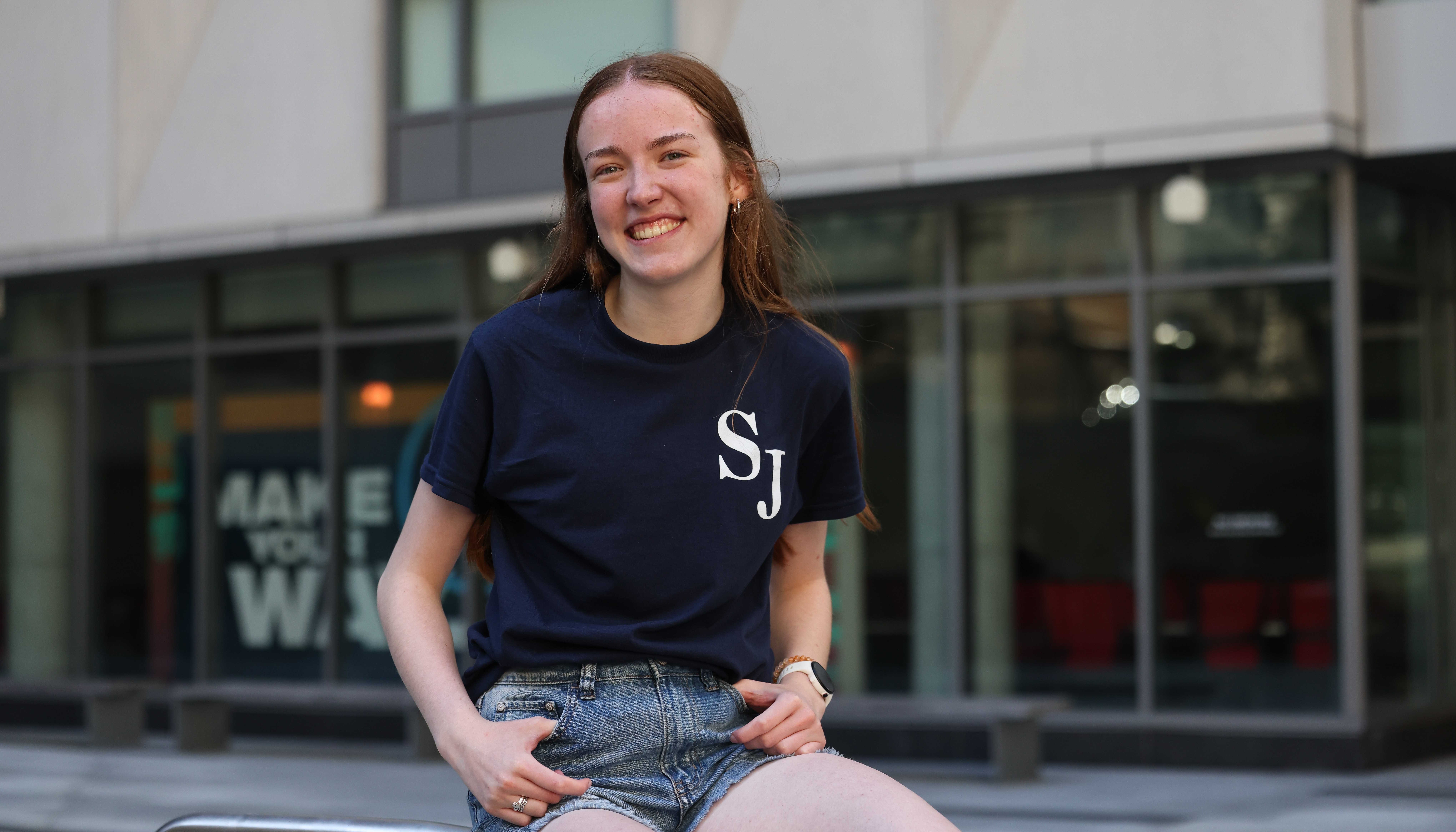 Maren Halpin smiles as she sits outside on the Suffolk campus wearing a Suffolk Journal shirt