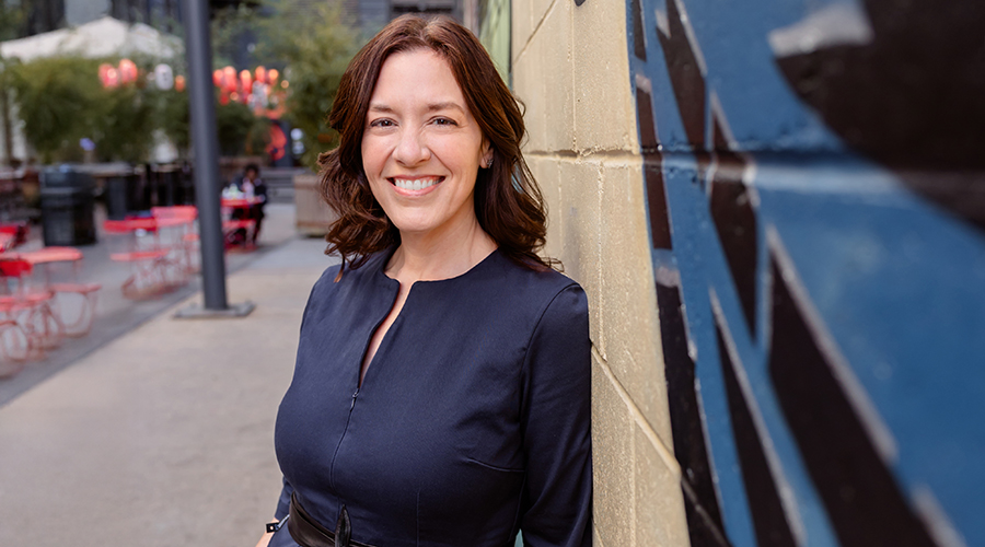 Mandi White-Ajmani leans against a concrete block wall with a city street behind her