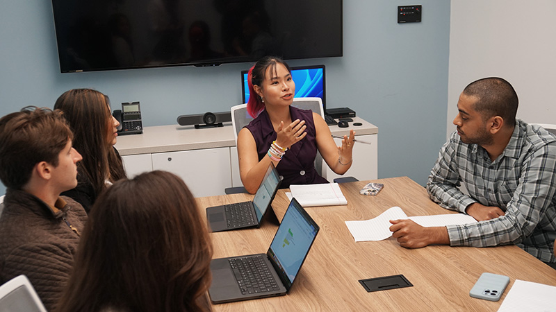 A group sit at a table listening to a colleague speak in a meeting space.