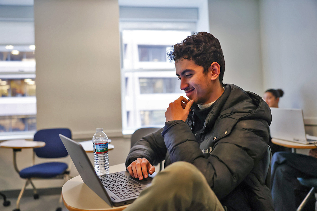 A Suffolk student sits casually at a desk in a computer science class with a laptop in front of him.