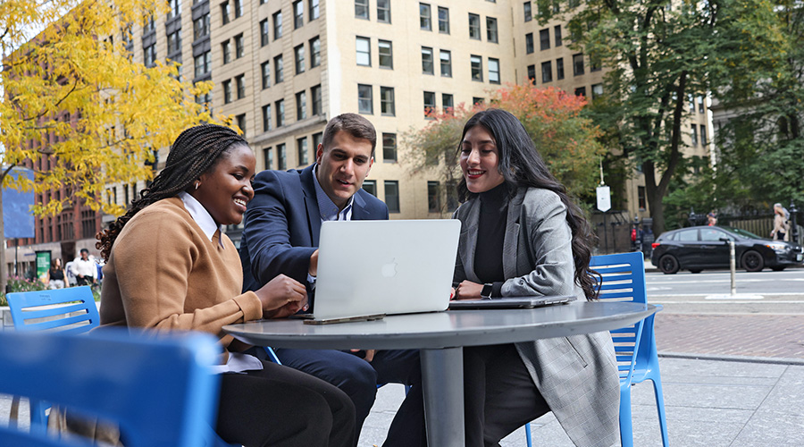 Suffolk Graduate students sit in discussion with a laptop at a table in a plaza adjacent to campus.