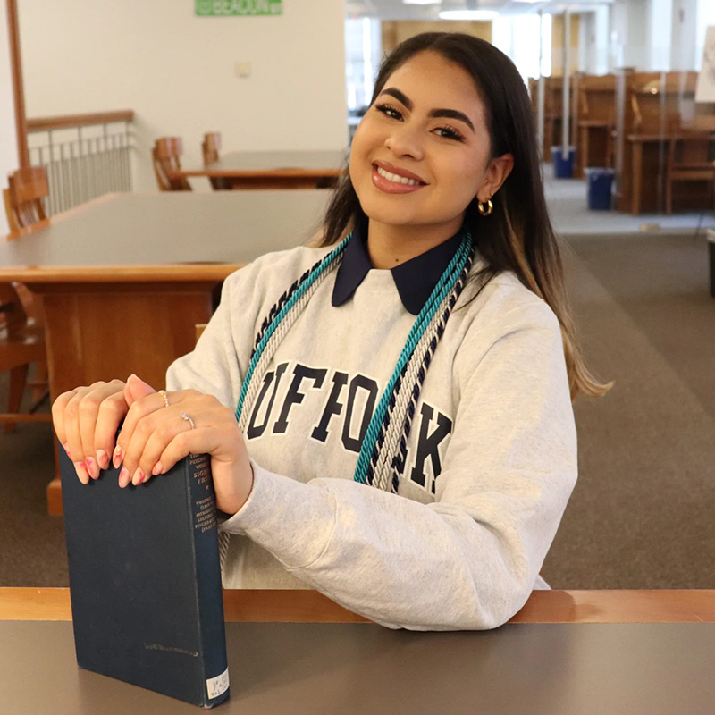 A student smiles in the library wearing her honors tassels and holding a book.