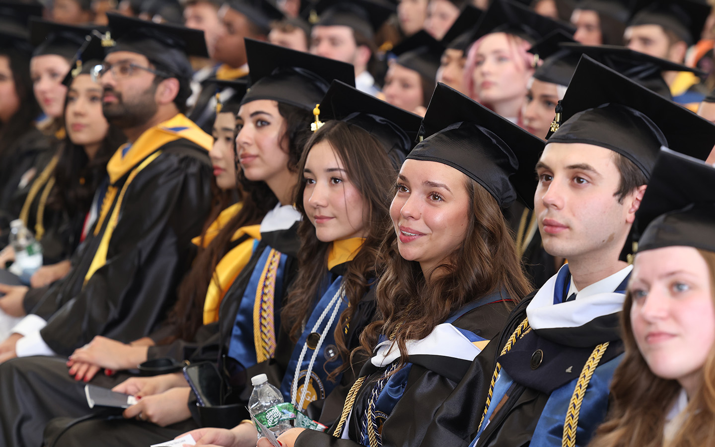 2026 graduates in caps and gowns smile as other graduates process at the 2026 Suffolk University College of Arts & Sciences Commencement Ceremony.