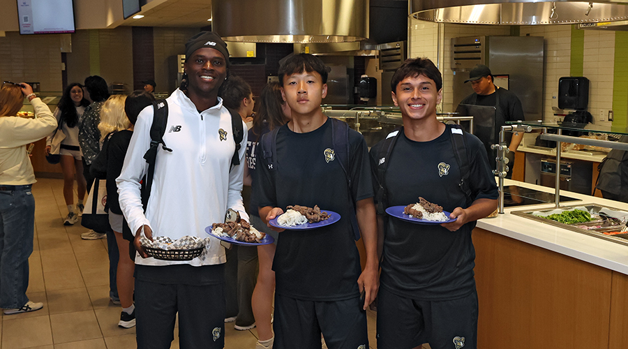 Three Suffolk students stand in the Samia Dining Hall holding plates of food.