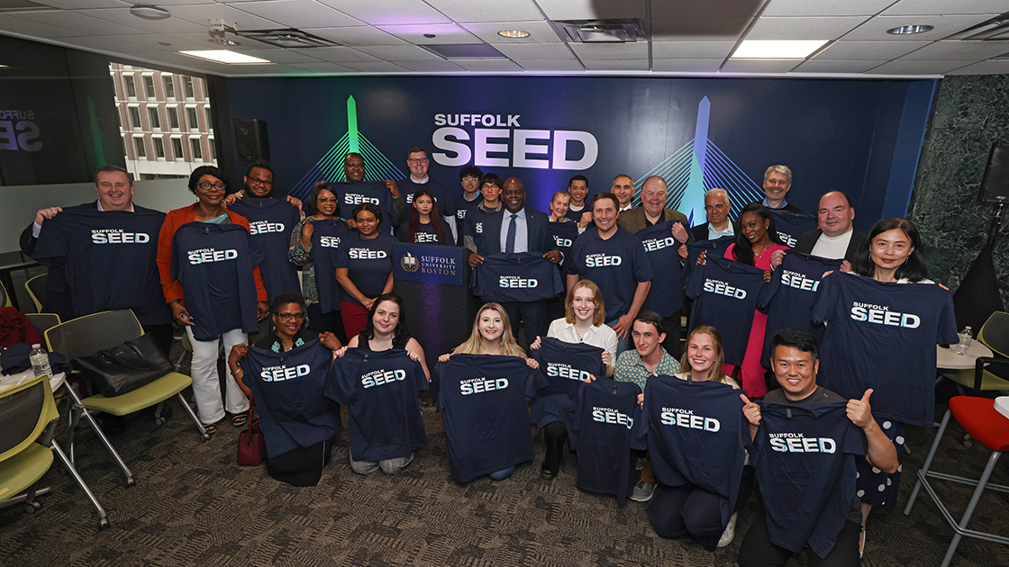 A group of Suffolk students and community members hold up Suffolk SEED shirts at a SEED event on campus.
