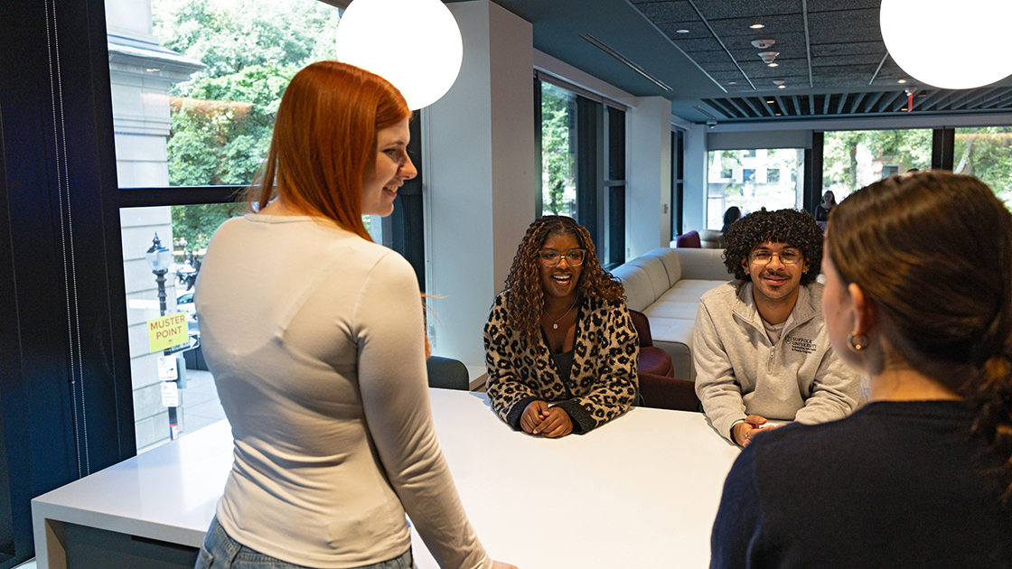 A small group of Suffolk students casually leaning against a table in discussion inside a lounge space in 101 Tremont, the newest residence hall on campus.