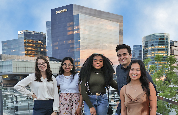 Five Suffolk students stand and pose together down near Boston's waterfront with city buildings behind them.