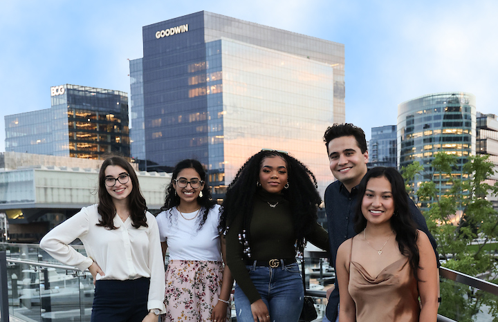Five Suffolk students stand and pose together down near Boston's waterfront with city buildings behind them.
