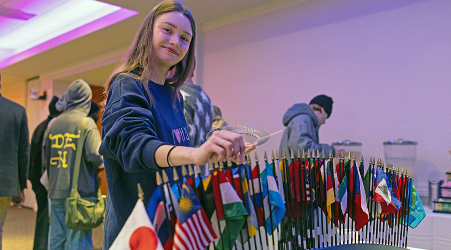A Suffolk student smiles standing behind a display of flags of the world at an International students event on campus.